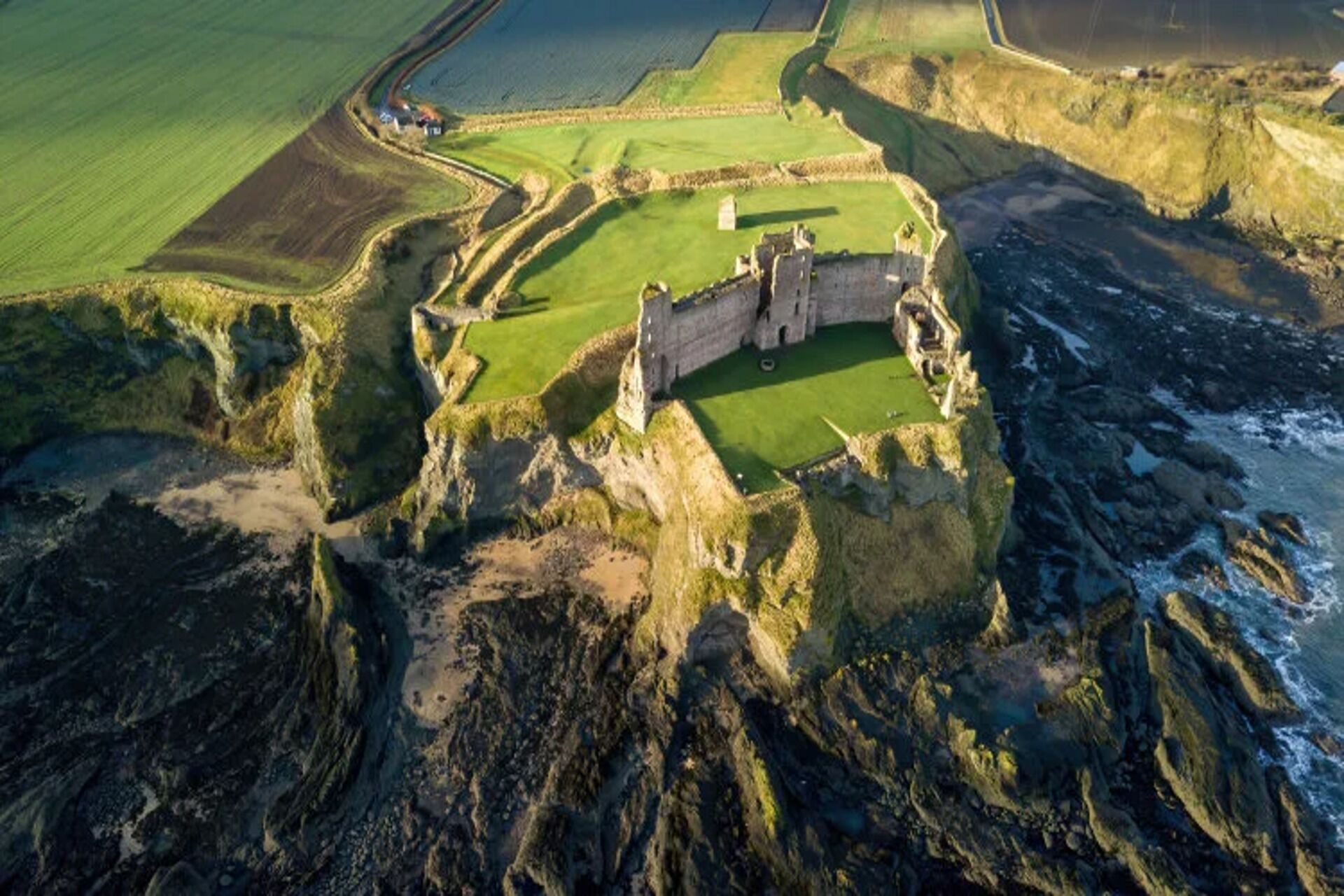 Una vista aerea di un castello circondato da campi verdi e una costa.