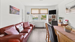 A cozy living room with a leather sofa, a dining table with chairs, a TV, and a window with blinds.