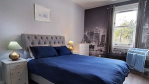 A bedroom with a large bed, two bedside tables with lamps, a cityscape mural, and a window with curtains.