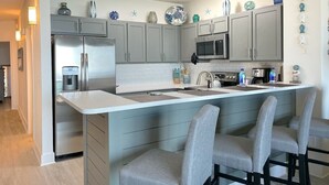 A modern kitchen with grey cabinets, a stainless steel refrigerator, a microwave, and a sink. There are bar stools and decorative items on the counter.