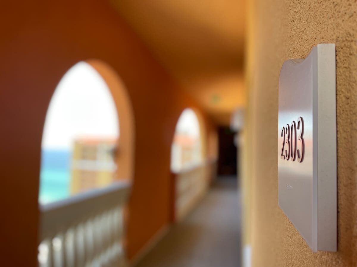A corridor with arched doorways leading to a balcony overlooking the sea. A sign with the number 2303 is visible on the wall.