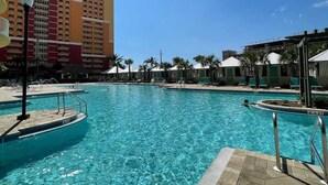 A swimming pool with clear blue water, surrounded by lounge chairs and umbrellas, with a hotel building in the background.