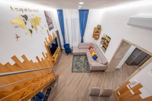 A modern living room with a wooden staircase, a sectional sofa, a blue armchair, a map on the wall, and a small bookshelf. - Monem's House-door (Milano)
