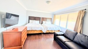 Hotellihuone, jossa on kaksi sänkyä, televisio, puinen lipasto, nahkasohva ja suuri ikkuna, jossa on verhot.