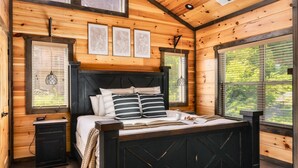 Un dormitorio de cabaña de madera con una cama grande, una mesita de noche y una ventana con persianas.