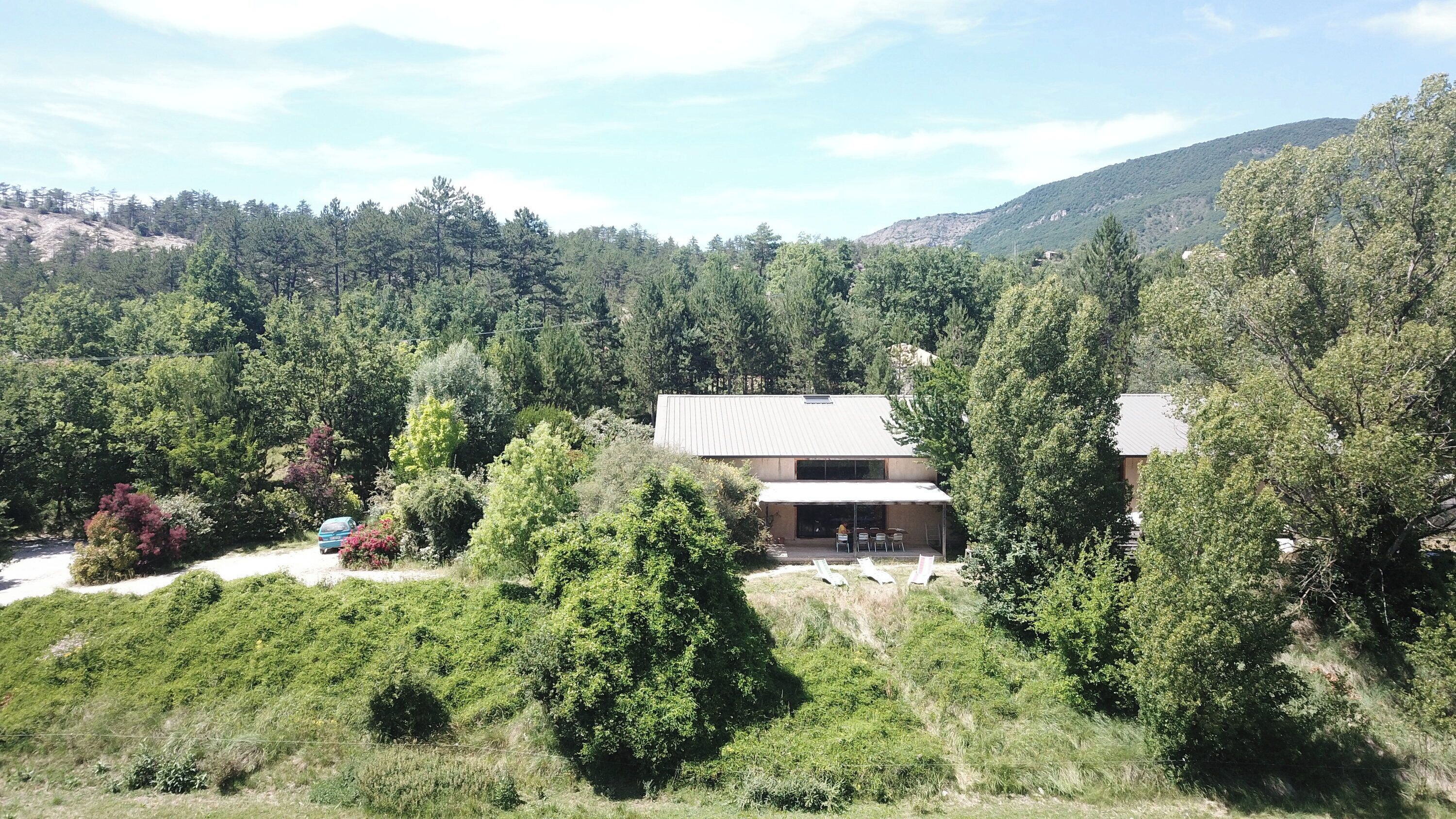 A modern house surrounded by dense forest, with a visible patio area and a car parked nearby.