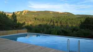 Piscina con cubierta de madera y escalera metálica, rodeada de bosque y montañas.