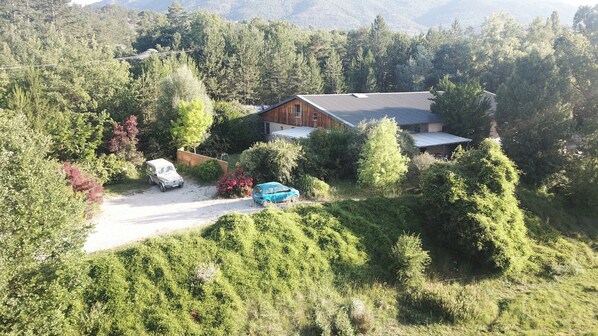 A cabin surrounded by dense forest, with a gravel driveway and two parked vehicles.