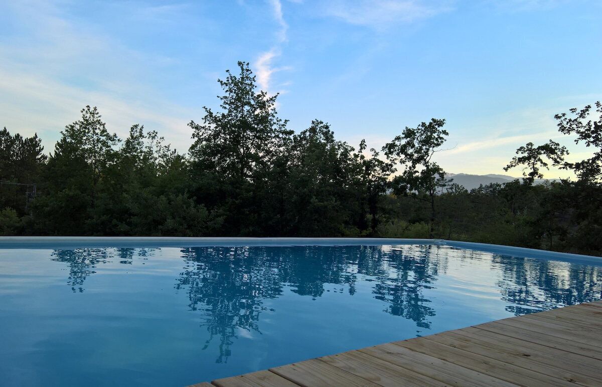 En pool med kristallklart vatten och en träbrygga runt om, omgiven av träd och en klar himmel.