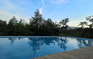 A clear swimming pool with a wooden deck, surrounded by trees and a clear sky.