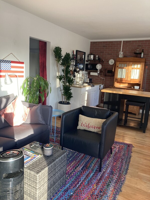 A living room with a black leather sofa, a colorful rug, and a coffee table. There is a kitchen area with a brick wall, a wooden cabinet, and various kitchen items.