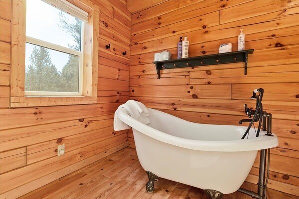 A wooden cabin bathroom with a freestanding bathtub, a window, and a shelf with toiletries.