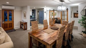 Un comedor con una mesa de madera y sillas de mimbre, una cocina al fondo y una sala de estar con un sofá y una planta.