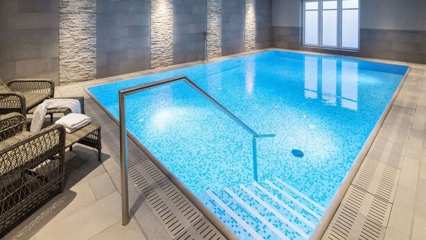 Una piscina interior con agua cristalina azul, rodeada de paredes de piedra y mobiliario de mimbre.
