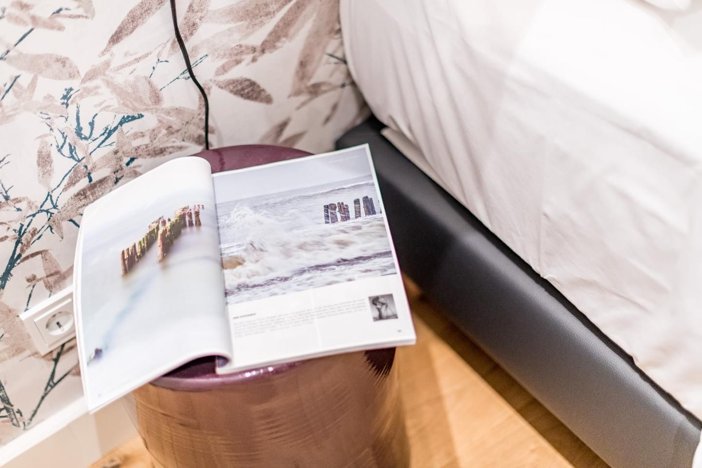 Un magazine est posé sur un tabouret en bois, à côté d’un lit dont la tête de lit est blanche.