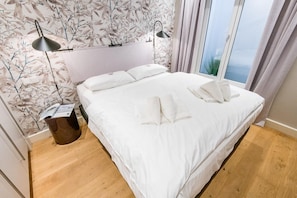 A neatly made bed with white linens and pillows, a modern lamp, and a window with curtains. - Tualighaker Hüs, Whg. 01 - Tualighaker Hüs (Sylt)