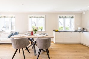 A modern kitchen with white cabinets, a dining table with chairs, and large windows with curtains. - Tualighaker Hüs, Whg. 01 - Tualighaker Hüs (Sylt)