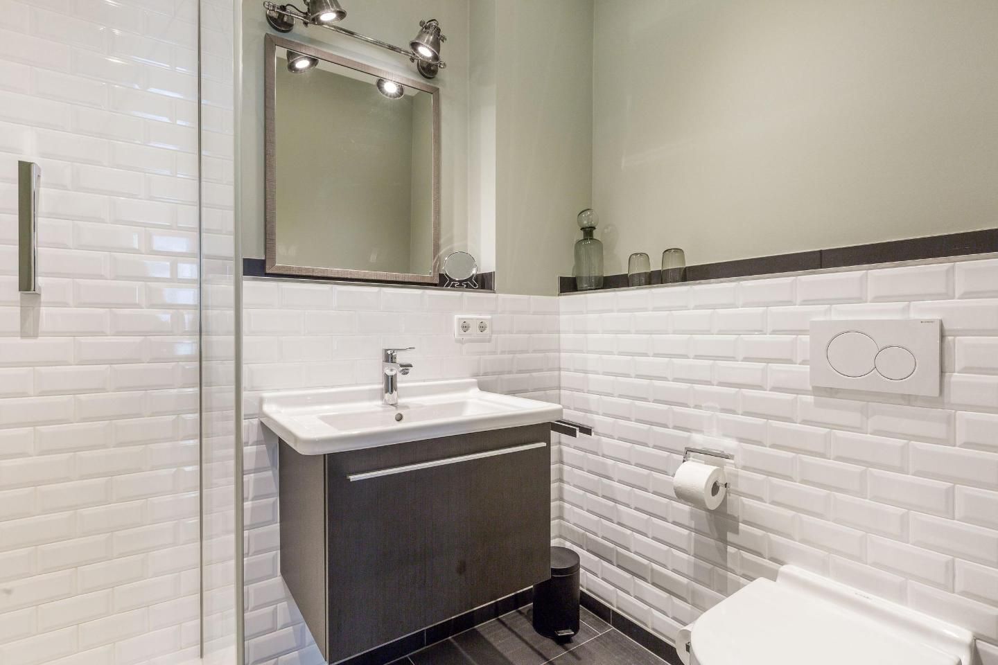Ein modernes Badezimmer mit weiß gefliestem Fliesenspiegel, einem weißen Waschbecken mit dunklem Unterschrank, einem Spiegel mit Deckenleuchten und einer Toilette mit Papierhandtuchspender.