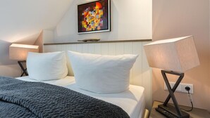 A hotel room with a bed, two bedside lamps, a painting on the wall, and a wooden shelf. - Art your Life - Residence, Whg. 9 Iliya - Art your Life - Residence Seeburg (Sylt)