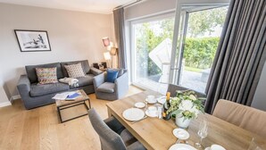 A modern living room with a grey sofa set, a wooden coffee table, and a dining area with a wooden table set for four. - Ringhoog-Hüs, Whg. 2 "Waldidyll" - Ringhoog Hüs (Sylt)