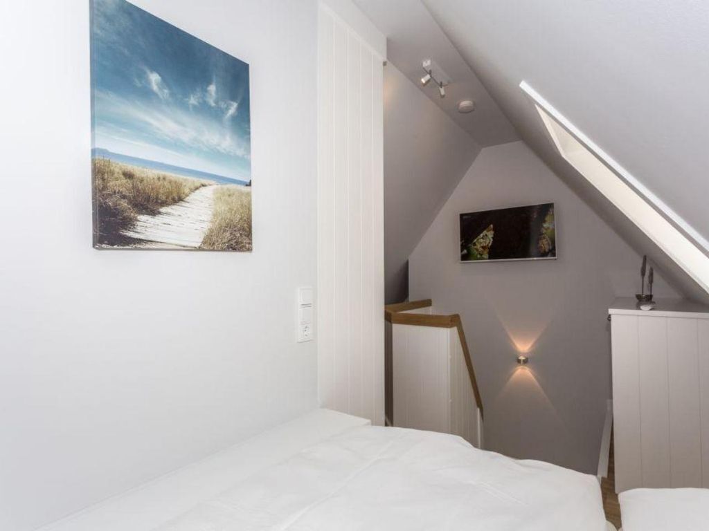 A bedroom with a white bed, a painting of a beach, a desk with a chair, and a sloped ceiling with recessed lighting.