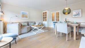A modern living room with a sofa, coffee table, dining table, and chairs. There is a kitchen area with a window and a pendant light. - House Nordmarkgarten, Whg. 5 - House Nordmarkgarten (Sylt)