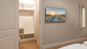 A bedroom with a wooden floor, a bed, a painting of a sunset over a beach, and a closet with shelves and a hanging rack. - House Nordmarkgarten, Whg. 5 - House Nordmarkgarten (Sylt)
