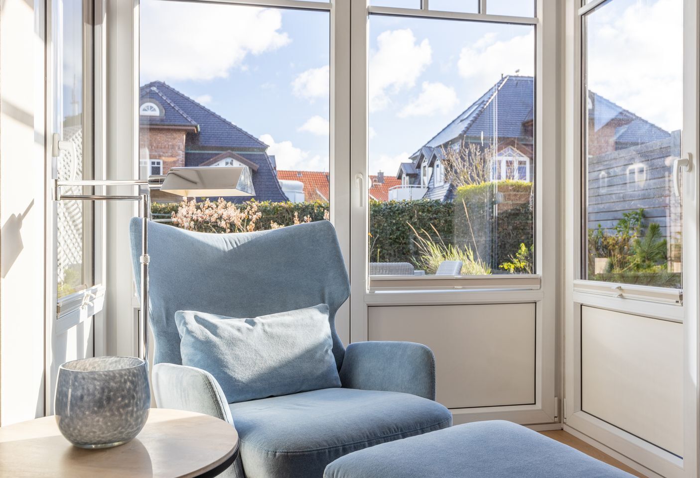 A modern living room with a blue armchair, a small table, and a view of neighboring houses through large windows.