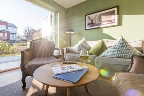 A cozy living room with a sofa, armchair, and coffee table. There are two glasses of wine and a book on the table. - Villa Maybach, Whg. 4 Grüner Wim - Villa Maybach (Sylt)