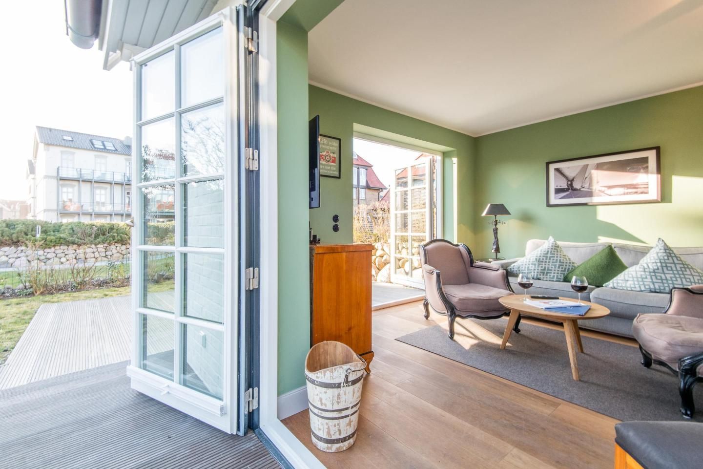 A spacious living room with a large sliding glass door leading to a garden, a sofa set, a coffee table, and a small wooden cabinet.