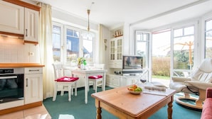 Eine helle Küche mit weißen Schränken, ein Essbereich mit Tisch und Stühlen, ein Fernseher und ein Blick ins Freie durch große Fenster.