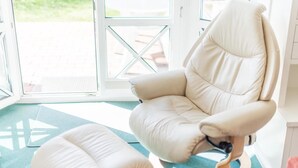Ein Zimmer mit einem weißen, gepolsterten Sessel und Hocker, Holzboden und Blick durch eine Glastür.