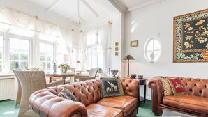 A well-lit room with a brown leather sofa, wicker chairs, a dining table with floral arrangements, and a large framed artwork on the wall. - Bädervilla, Whg. 28a - Bädervilla (Sylt)