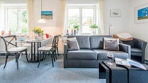 A modern living room with a grey sofa, a dining table with chairs, and a coffee table. There are two windows with curtains, a painting on the wall, and a lamp on a side table. - Bädervilla, Whg. 28b - Bädervilla (Sylt)