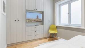 A bedroom with a bed, a TV mounted on a white wardrobe, a window with a view of a lighthouse, and a yellow chair.