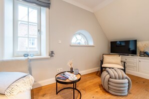 A cozy living room with a television, a comfortable chair, a small table with books, and a window with a view of a brick building.