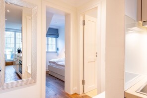 A narrow hallway with a bed, a window with blue curtains, and a kitchen area visible through an open door. - Maison Victor - Maison Victor, Apt. 4 (Sylt)