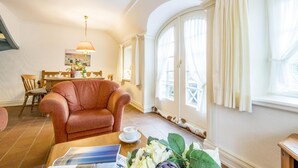 Ein gemütliches Wohnzimmer mit einem bequemen Sessel, einem Esstisch mit Stühlen und einem Fenster mit Vorhängen.