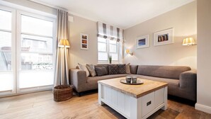A modern living room with a grey sofa, a wooden coffee table, and a large window with curtains. - House Steinmannstraße 77, half of the house A (Sylt)