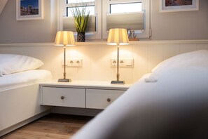 A hotel room with two beds, a nightstand, two lamps, a potted plant, and a window with blinds. - House at Steinmannstraße 77, half of House A (Sylt)
