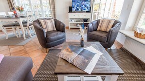 Un salon comprenant deux fauteuils en cuir, une table basse avec des magazines, un téléviseur diffusant une vue sur la ville, et un coin repas avec une table et des chaises blanches.