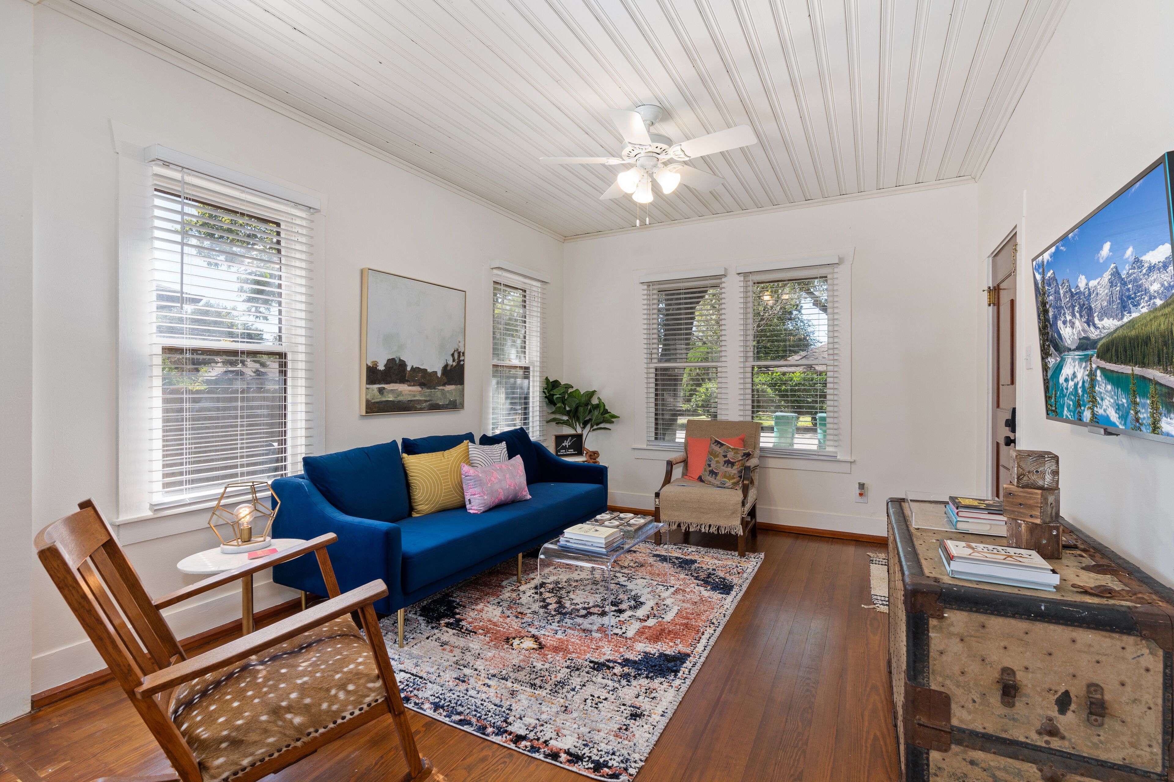 A living room with a blue sofa, wooden chairs, a patterned rug, and a ceiling fan.
