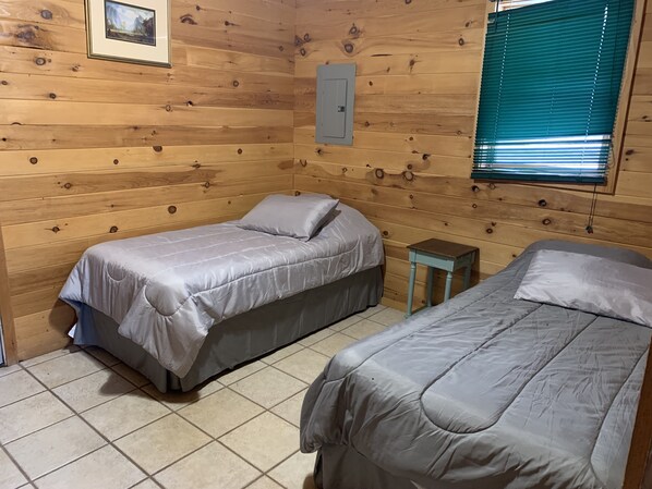A cabin-style room with two beds, wooden walls, a small table, and a window with blinds.