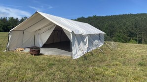 Una gran tenda blanca amb teulada plana, instal·lada en un camp verd amb un turó forestal al fons.