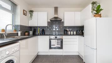 A compact kitchen with white cabinets, a stainless steel range hood, a built-in oven, a washing machine, and a small countertop with various appliances and plants.