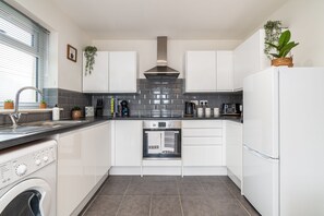 A compact kitchen with white cabinets, a stainless steel range hood, a built-in oven, a washing machine, and a small countertop with various appliances and plants. - Stylish Southampton Abode (Freemantle)