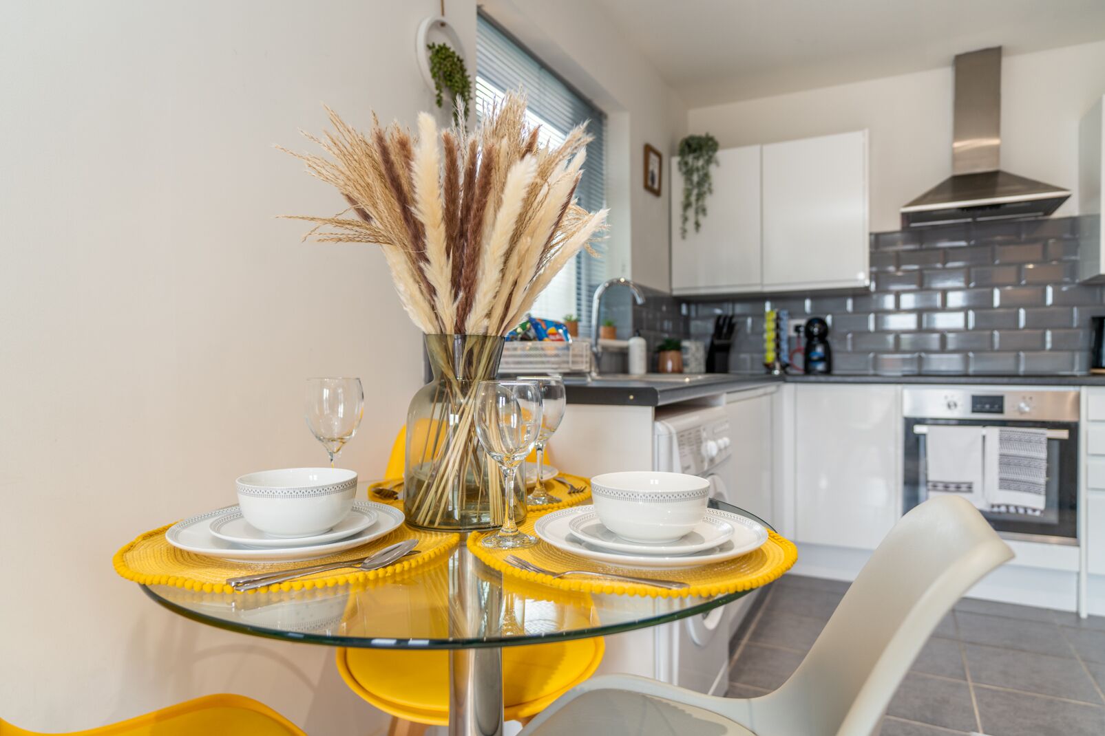 A modern kitchen with a glass dining table, white chairs, and a vase with pampas grass.