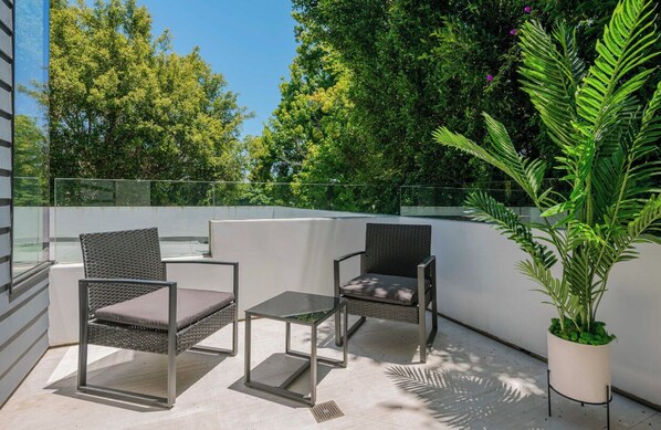 A balcony with two wicker chairs and a small table, surrounded by lush greenery and a clear sky.