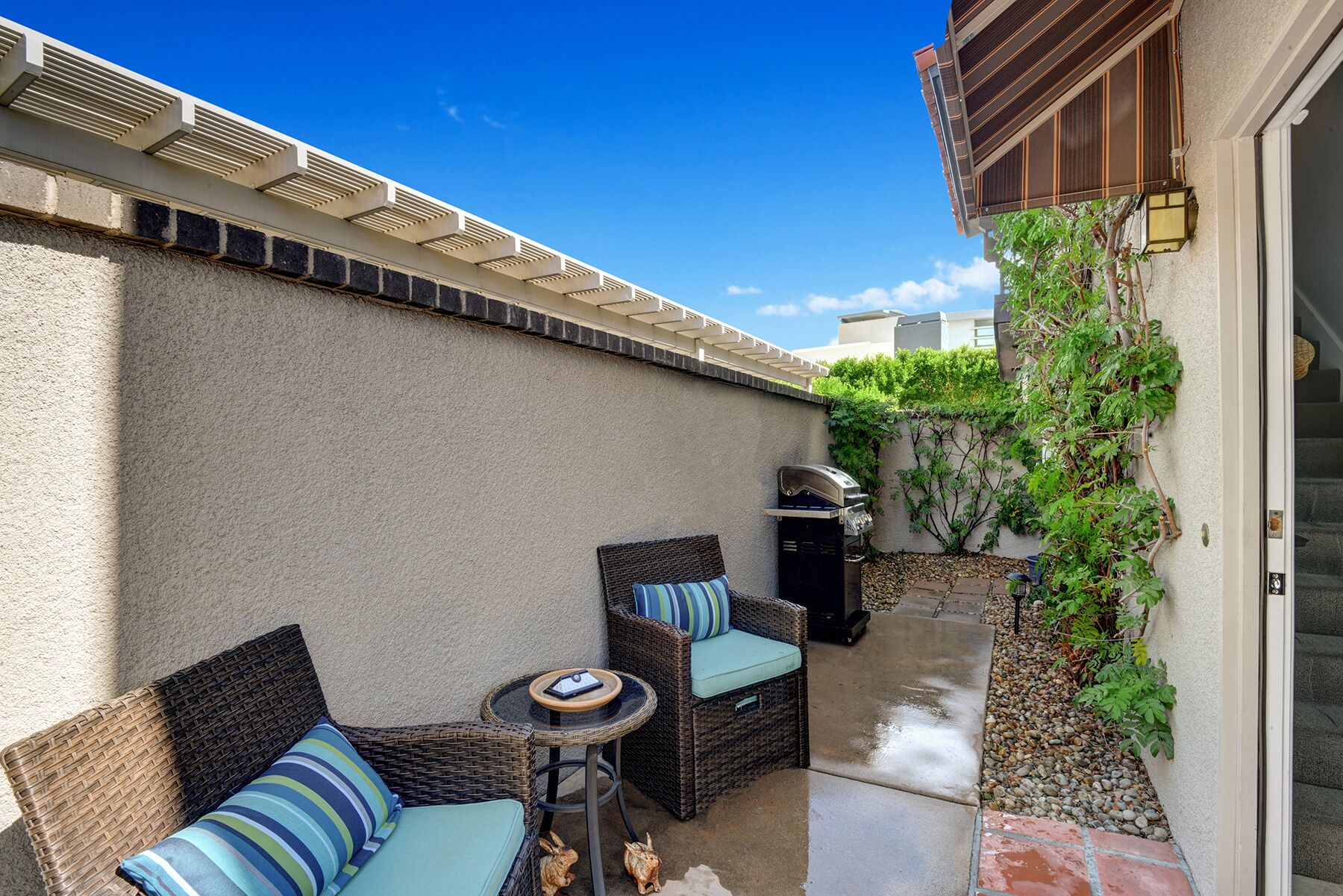 Une terrasse dotée de meubles en osier et d’un barbecue, entourée d’un mur de pierre et de verdure.
