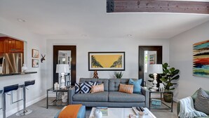 Una sala de estar moderna con un sofá gris, una mesa de centro y un reposapiés, un área de cocina con un refrigerador de acero inoxidable y un cuadro abstracto de gran tamaño en la pared.